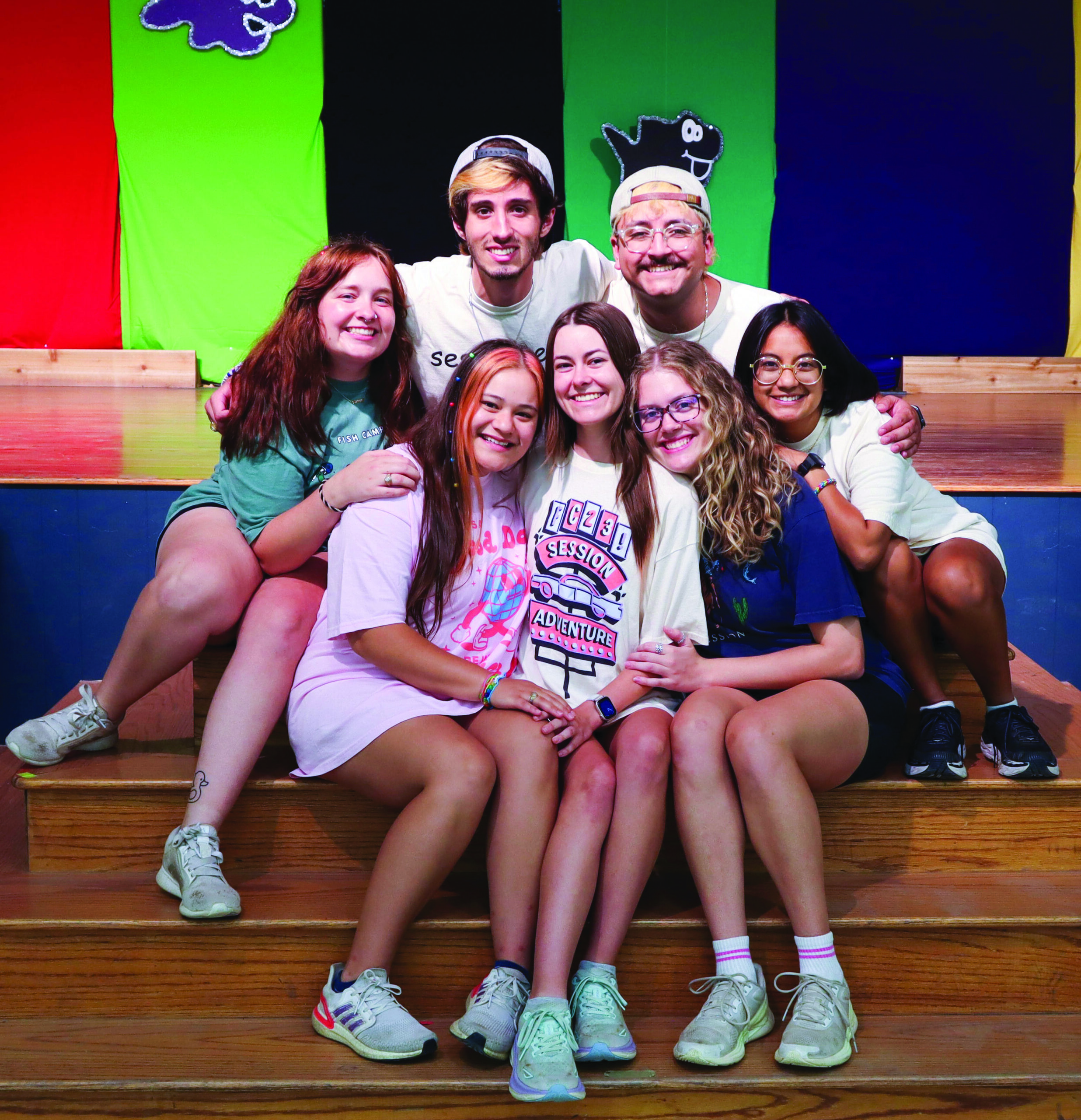 A group of college students are sitting on the stairs of a wooden stage and huddled together and smiling.