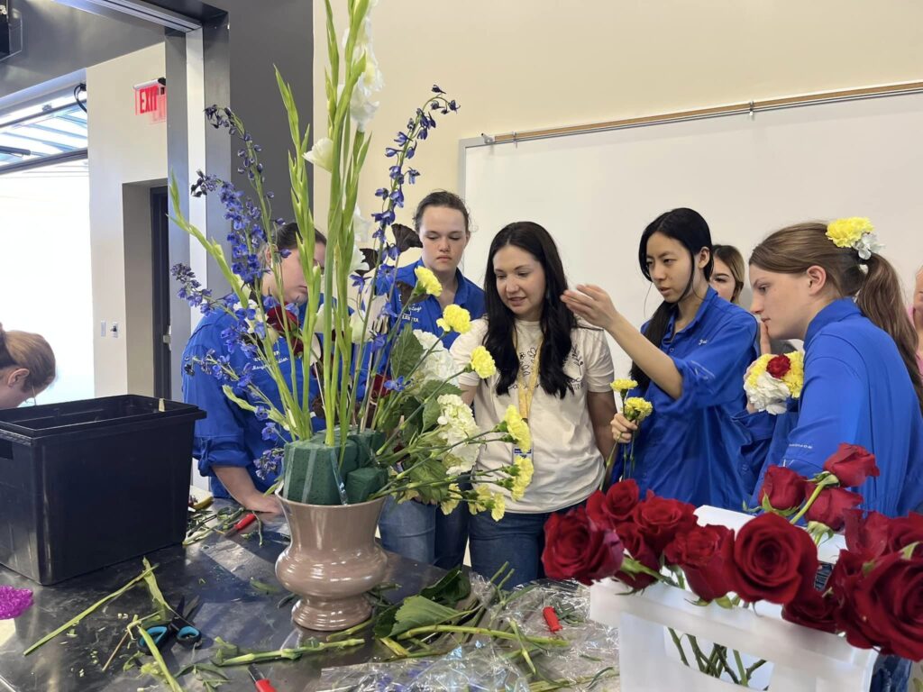 A young woman with long dark hair instructs her students on floral design. A large floral display is in front of them.