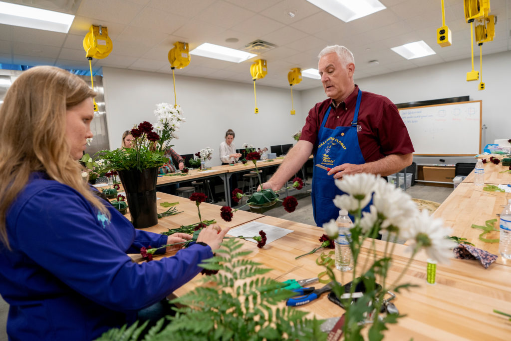 Professor handing a floral design to a student sitting behind a desk