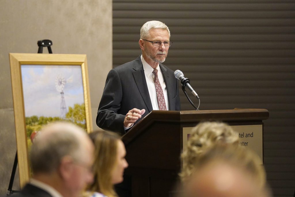 Dr. Jim Mazurkiwiecz talking at podium with painting on display next to him