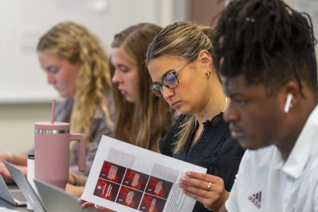 A line of students sitting at a table looking at their laptops, with one looking at a piece of paper