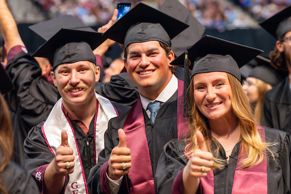 Graduating Aggies at a graduation ceremony posing with their thumbs up in the Gig 'Em gesture.